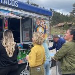 People stand in line at a colorful taco food truck in an outdoor setting, waiting to place their orders.