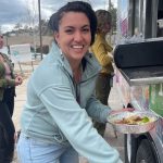 A woman in a blue jacket and jeans smiles while holding a container of food at a food truck. Other people are visible in the background.