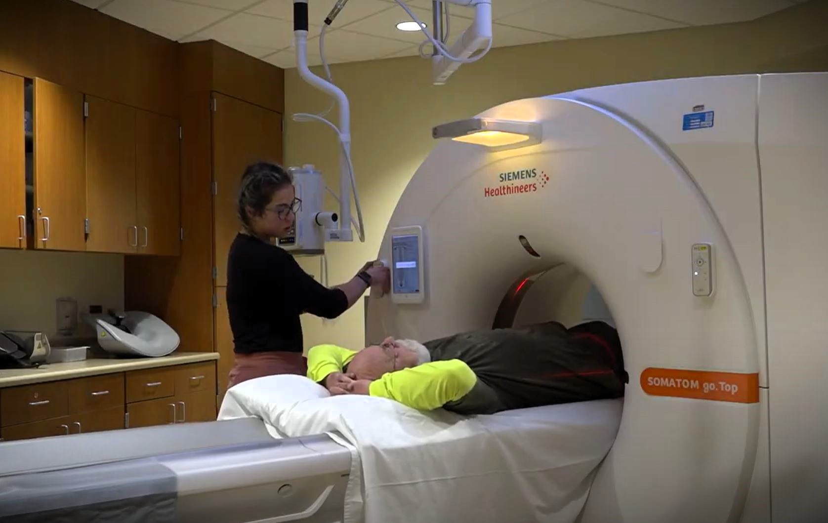 A medical technician assists an elderly patient lying on a bed as they undergo a CT scan in a hospital room. The patient is inside a Siemens Healthineers SOMATOM go.Top scanner.