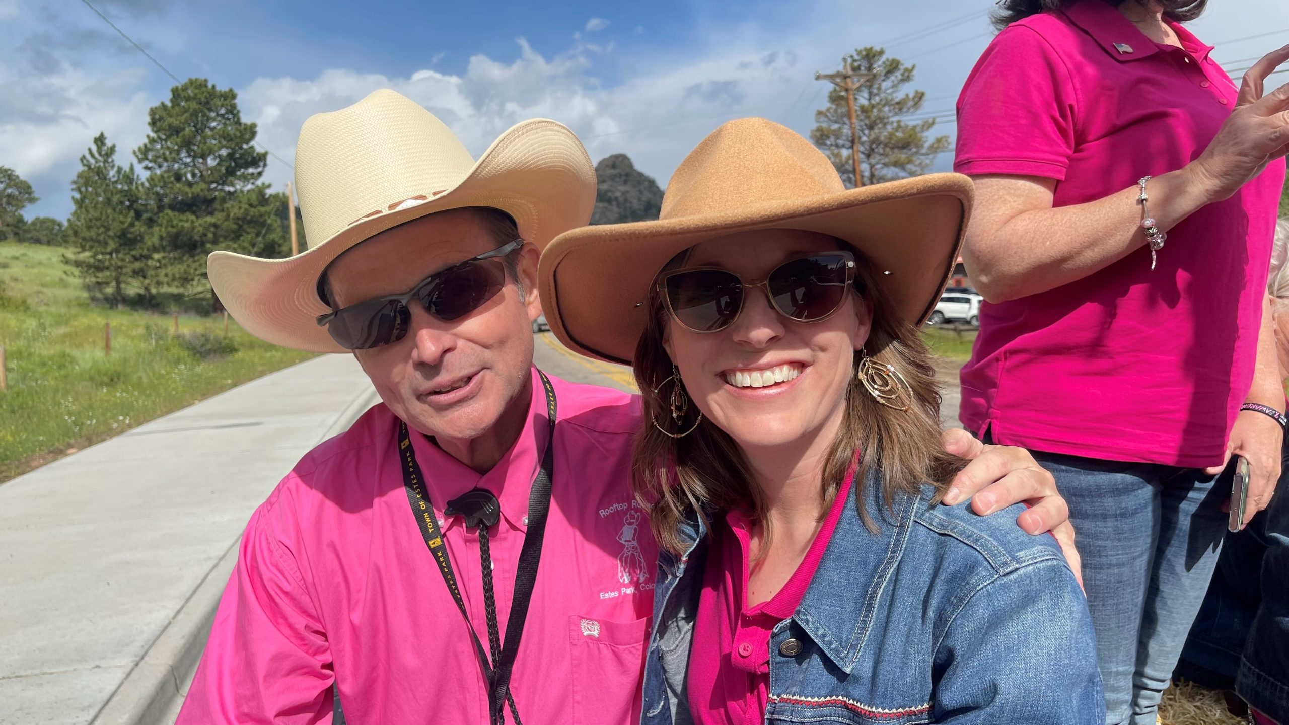 Two people wearing sunglasses and cowboy hats, both smiling and wearing pink shirts with black lanyards. They are outdoors on a sunny day.