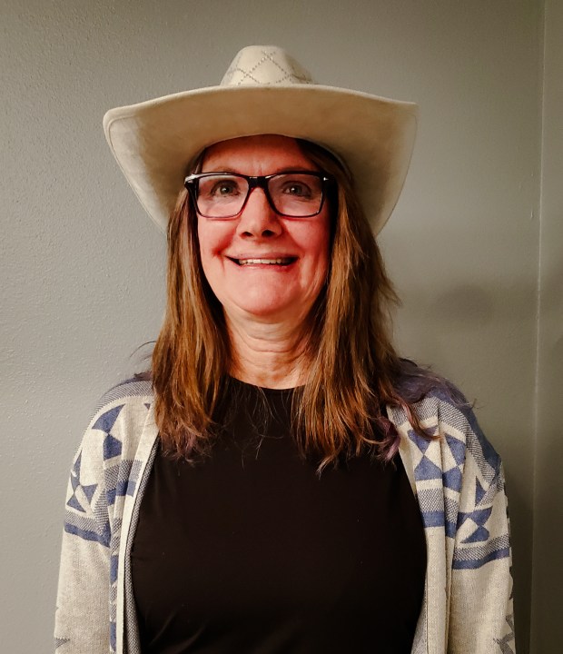 A woman with glasses and long hair smiles while wearing a white cowboy hat and a patterned cardigan over a black shirt. She is standing against a plain, light-colored wall.