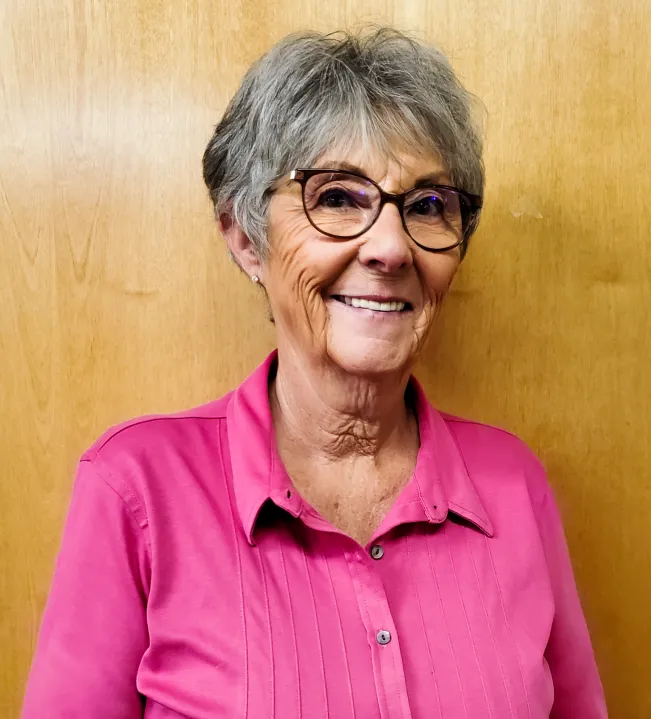 An older woman with short gray hair and glasses smiles while wearing a bright pink blouse. She stands against a wooden background.