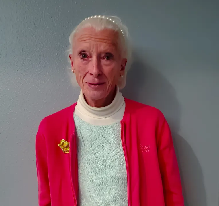An elderly person with white hair stands against a gray wall, wearing a red cardigan and a light blue shirt, with a yellow pin on the cardigan.