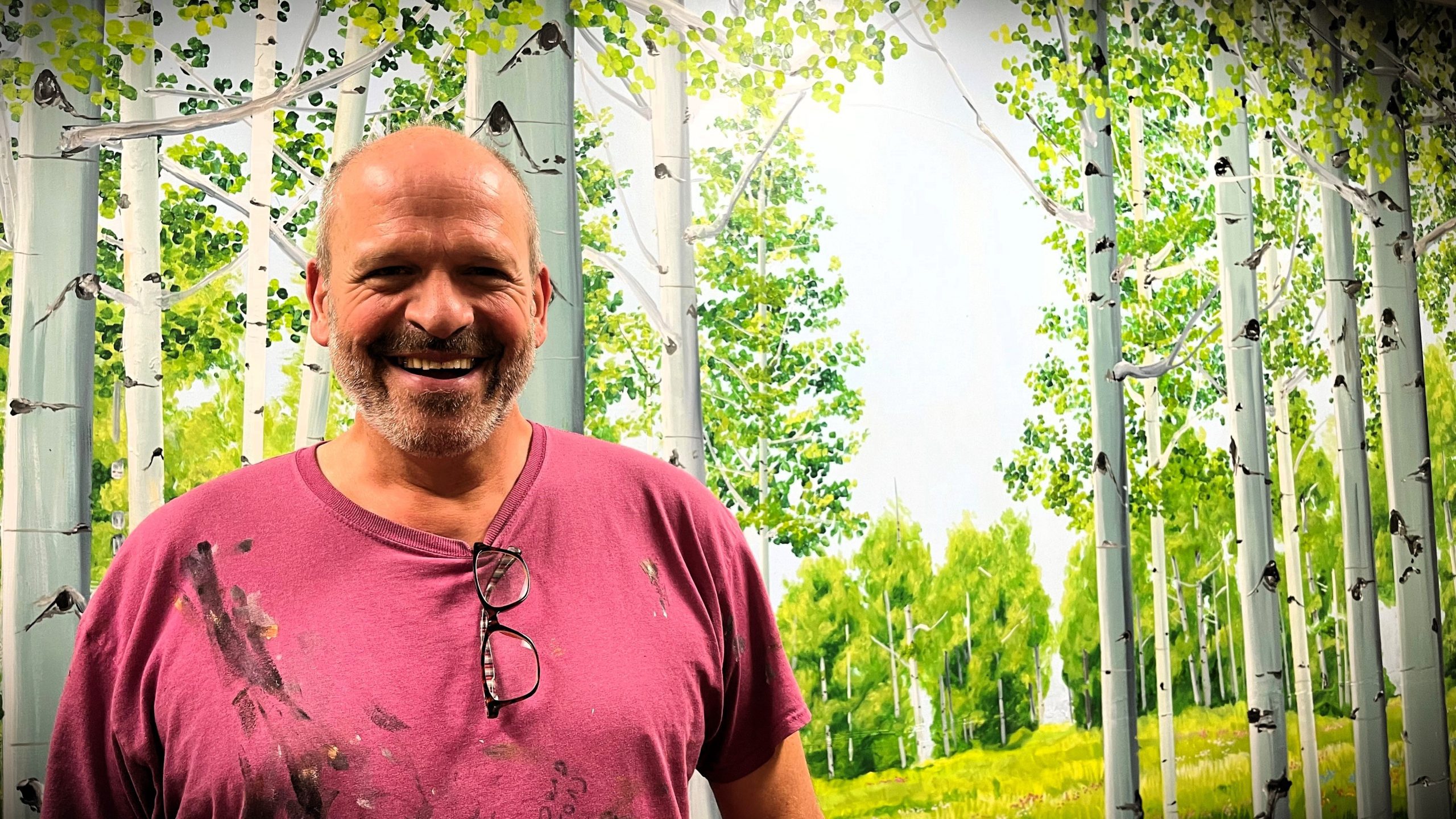 A man with a bald head and beard smiles while wearing a paint-stained pink shirt, standing in front of a detailed mural depicting a forest scene with birch trees and greenery.