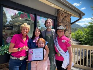 A group of five people, including a child, stands outside a building. One person holds a "Pinkiest Pink" award certificate for "Bear & Bee.” Another person is wearing a sash and cowboy hat.