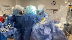 Three medical professionals wearing blue surgical gowns and protective gear perform a procedure in a sterile operating room. A clock on the wall shows the time and medical equipment is seen around.