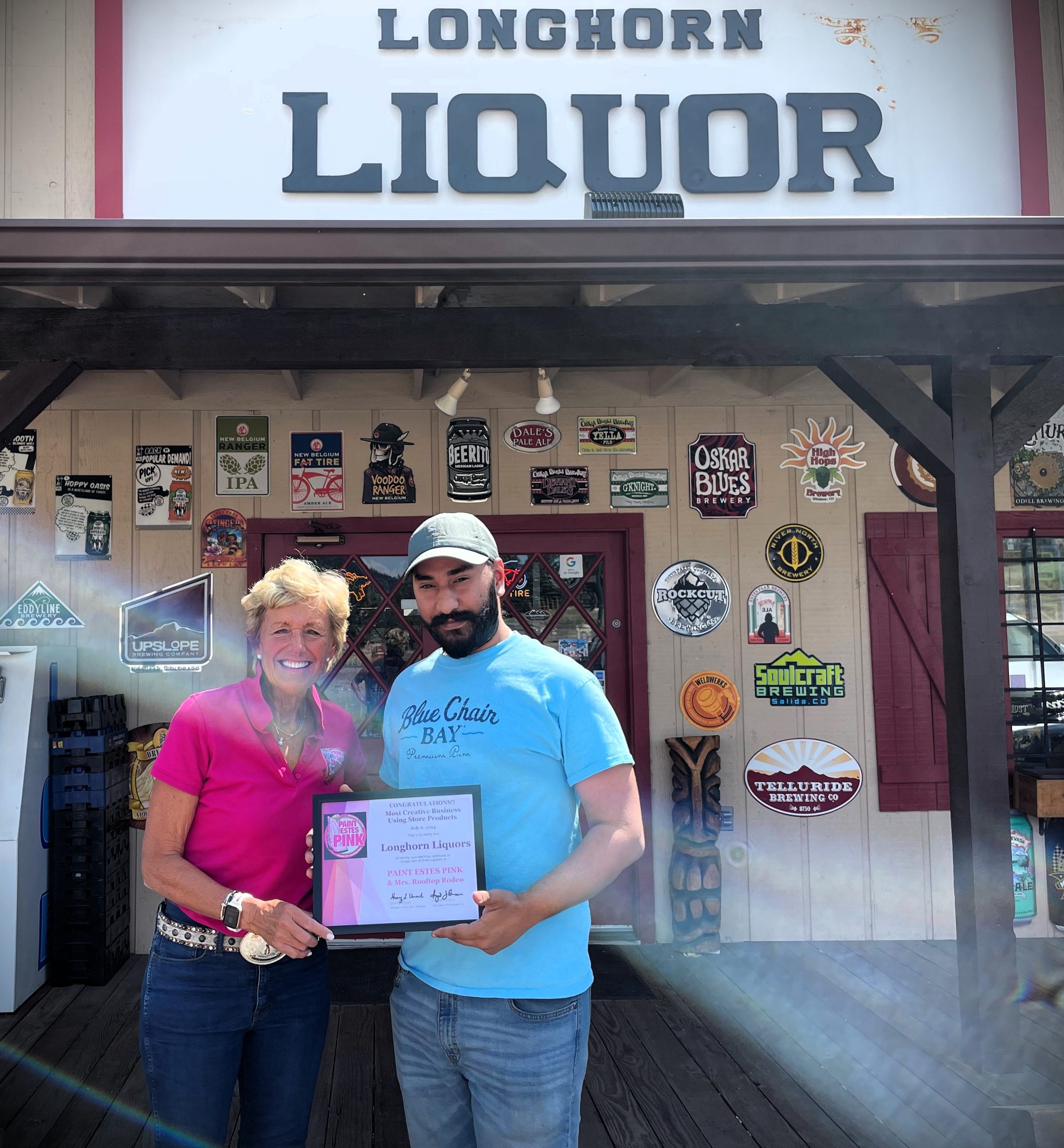 Two individuals stand outside Longhorn Liquor. One holds a certificate of recognition. The store is adorned with various beer and beverage signs.