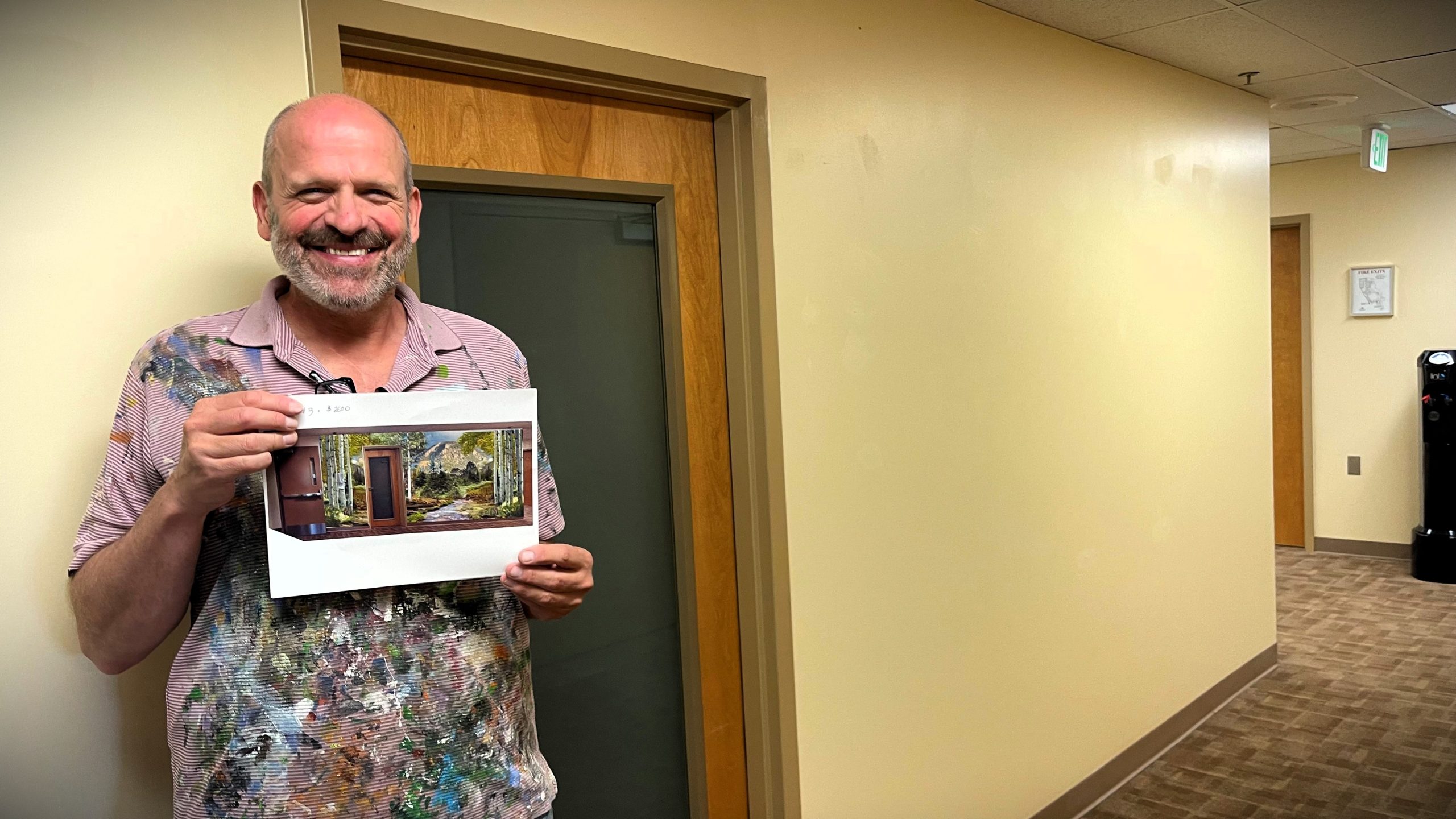 Man standing in a hallway holding a printed image of a room. He is smiling and wearing a paint-stained shirt.