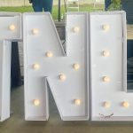 Large illuminated letters "TNL" displayed outside with light bulbs embedded in them, set against a rustic wooden table and plants.