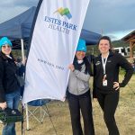 Three individuals stand outdoors next to an Estes Park Health banner; one holds the banner. They are dressed in casual attire with beanies and are smiling, with a small gathering in the background.