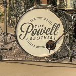 Close-up of a drum kit with a white bass drum displaying the text "The Powell Brothers." The drum kit is set up on an outdoor stage with various wires and microphones around it.