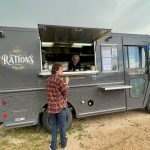 A food truck labeled "Rations" serves a customer in a plaid shirt. The truck is parked on a dirt area near a marina.