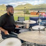 A person smiles while playing drums outside near a community center with mountains in the background. Nearby are other musical instruments and a computer.