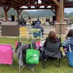 Audience members sitting on lawn chairs watch a live band performance under a wooden pavilion in a scenic outdoor setting with mountains in the background.