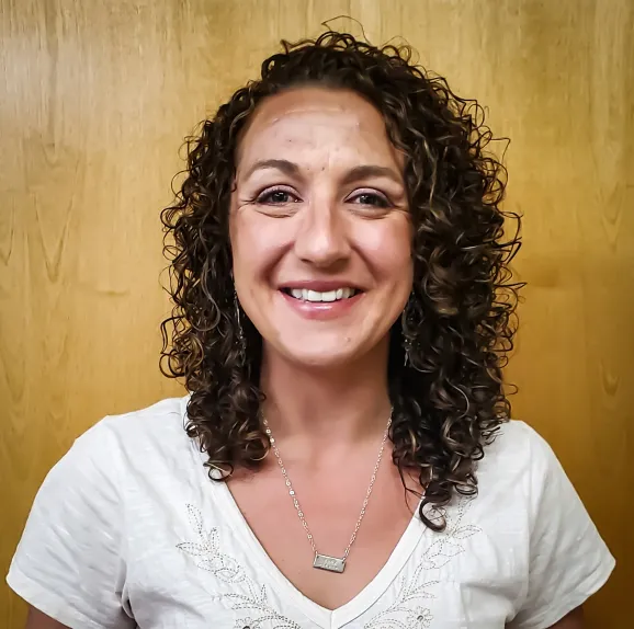 A woman with curly brown hair smiles at the camera, standing in front of a wooden background. She is wearing a white top and a necklace.