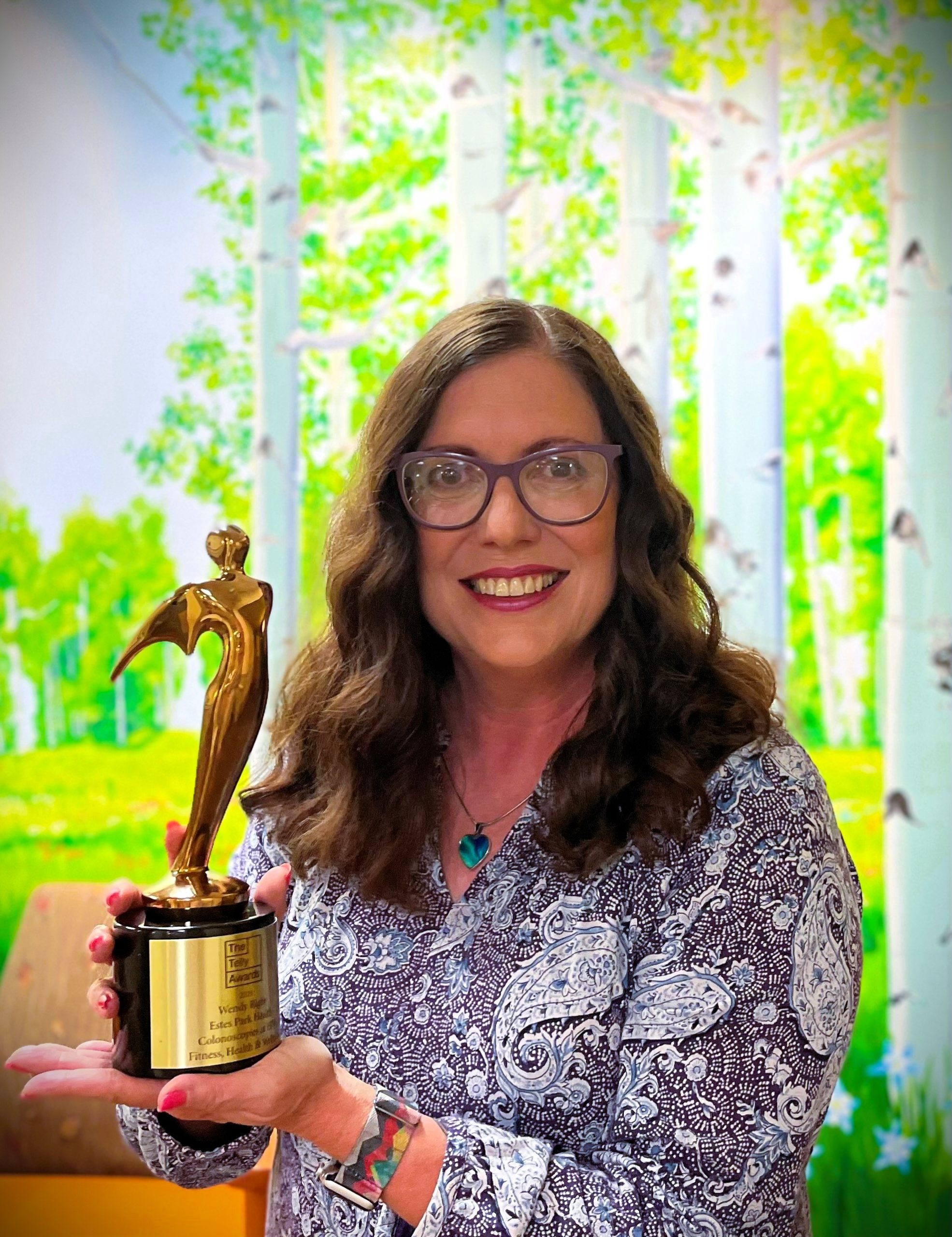 A woman with glasses and long brown hair, wearing a patterned blouse, smiles while holding a golden trophy. A colorful backdrop of trees and greenery is visible behind her.
