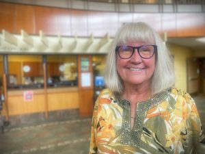 A woman with long gray hair and glasses, wearing a floral blouse, stands smiling in front of a reception area inside a building.