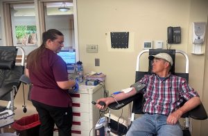 A man wearing a plaid shirt donates blood as a healthcare worker in a maroon uniform prepares equipment in a medical room with various devices and cabinets.