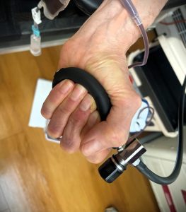 A person grips a black handle, with medical equipment attached to their hand and wrist, set against a background that includes a wooden floor and various medical devices.