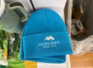 A blue beanie with "Estes Park Health" and a mountain logo embroidered on the front is placed on a surface. Objects in the background are blurred.