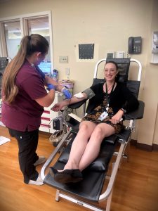 A woman is seated in a chair donating blood. A nurse in maroon scrubs is attending to her, placing a blood pressure cuff on her arm. The room has medical equipment on the walls and a tiled floor.