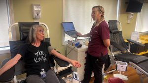 An elderly woman donates blood while seated in a donation chair, engaging in conversation with a healthcare worker who is inputting data into a laptop.