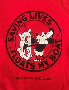 Red shirt with a cartoon mouse steering a ship's wheel. Text reads, "SAVING LIVES FLOATS MY BOAT." "GARTH ENGLUND BLOOD CENTER" is printed below.