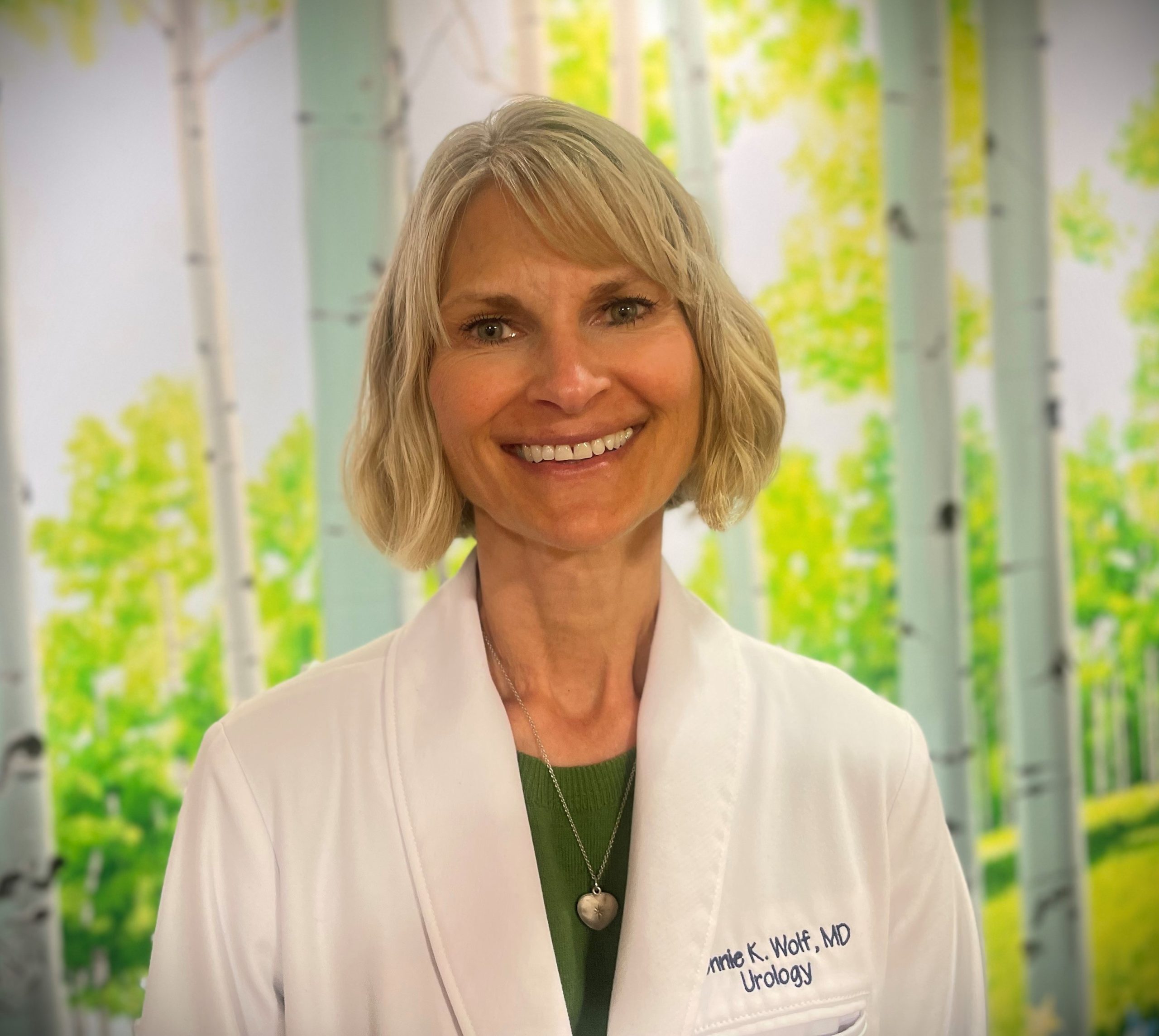 A smiling woman in a white lab coat stands in front of a background with white birch trees and green foliage.