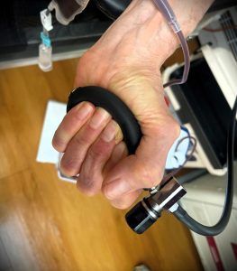 A close-up of a hand gripping a black stress ball with medical tubing attached to the wrist, taken on a wooden floor background.