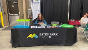 A woman sits behind an Estes Park Health booth displaying water bottles, brochures, pens, and t-shirts at an event.