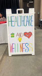 A sign with colorful lettering reads "Healthcare and Wellness" with related drawings including an apple, heart, and light bulb. A person sits at a table in the background.