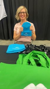 A woman with glasses and a light pink shirt sits at a table holding a blue beanie. Green tote bags and blue beanies are displayed in front of her.