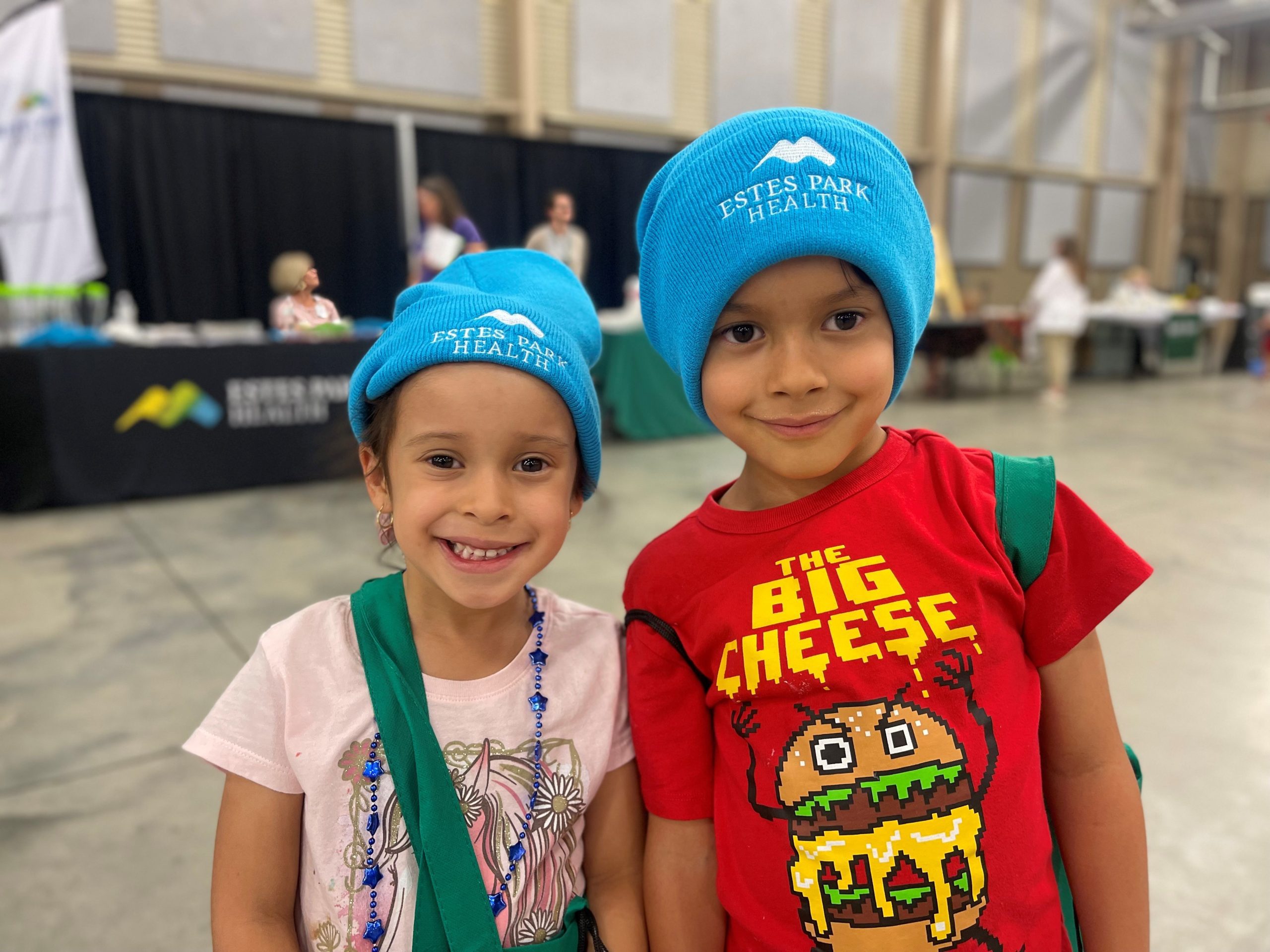 Two children wearing blue beanies and colorful shirts stand together indoors, smiling at the camera. A booth and several people are visible in the background.