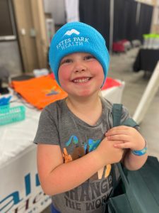 A smiling child wearing a blue "Estes Park Health" beanie holds a green bag. They are standing in what appears to be an indoor event with tables and banners in the background.