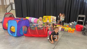 Several young children play in a colorful indoor play area with a toy tunnel, play mats, and large blocks, while a woman watches and interacts with them.