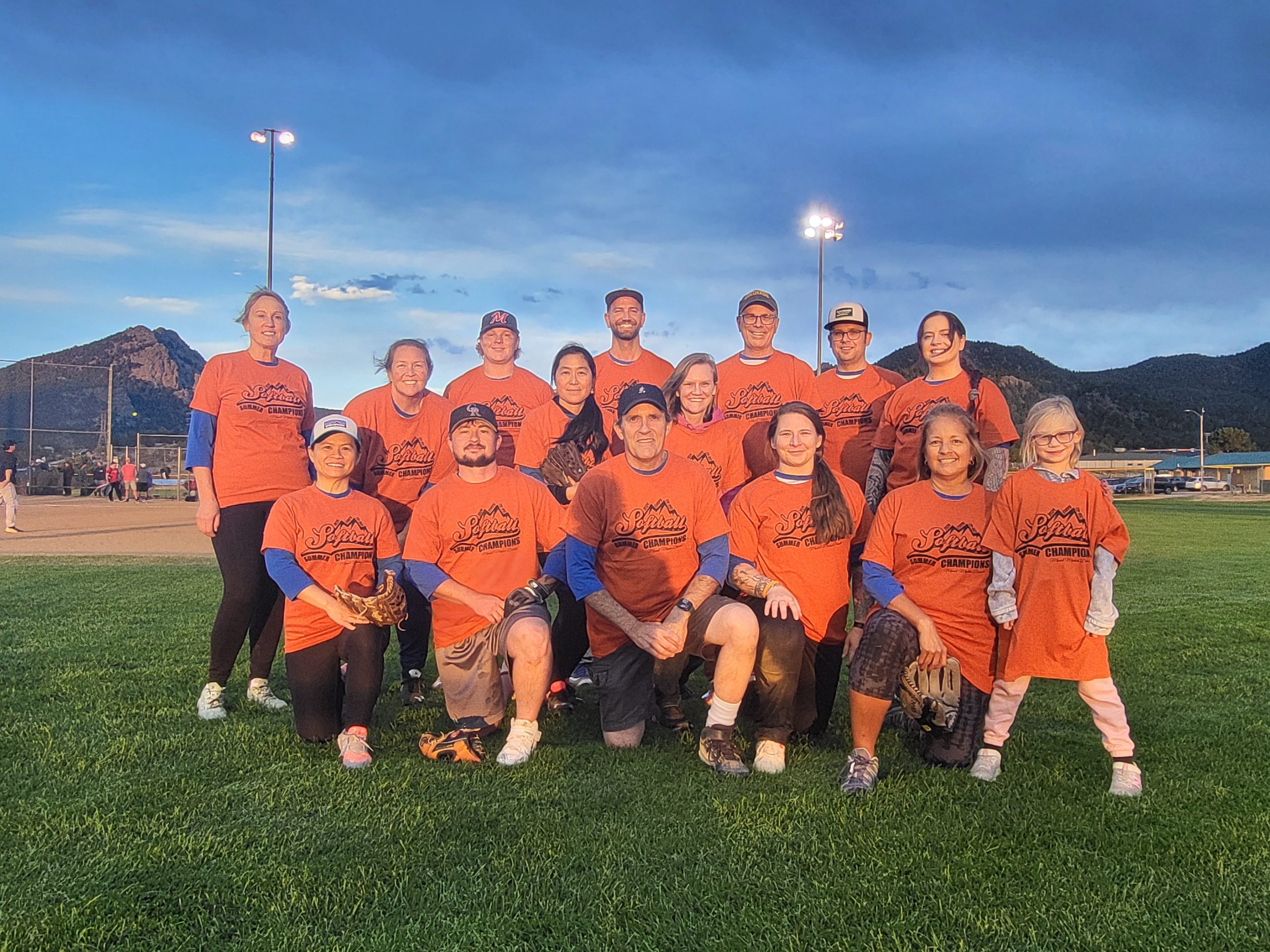 A group of 15 people wearing matching orange shirts poses for a photo on a grassy field with mountains and a cloudy sky in the background.