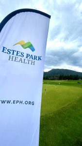 A banner with "Estes Park Health" and a logo stands near a golf putting green. The backdrop features cloudy skies and a mountainous landscape. The website www.eph.org is visible at the bottom.