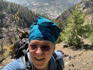 A person wearing a blue bandana and sunglasses stands on a rocky, mountainous trail with a town and forested landscape in the background. They have a large backpack on their shoulders.