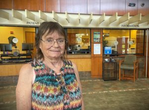 An older woman with glasses stands in a hospital lobby in front of signs reading "Patient Check-In" and "Information.