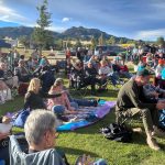 A crowd of people, some with dogs, are seated on chairs and blankets on a grassy area at an outdoor event with mountains in the background.