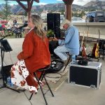 Two musicians, a woman and a man, perform on an outdoor stage. The woman is singing into a microphone, and the man is playing a guitar. Various musical equipment surrounds them. An audience is visible in the background.