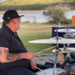 A person with tattoos and glasses plays the drums outdoors by a lake, wearing a black shirt and black cap. The background includes greenery and a body of water.