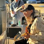 A seated man in a cap and glasses plays an electric guitar outdoors, illuminated by sunlight. Musicians and equipment are visible in the background.