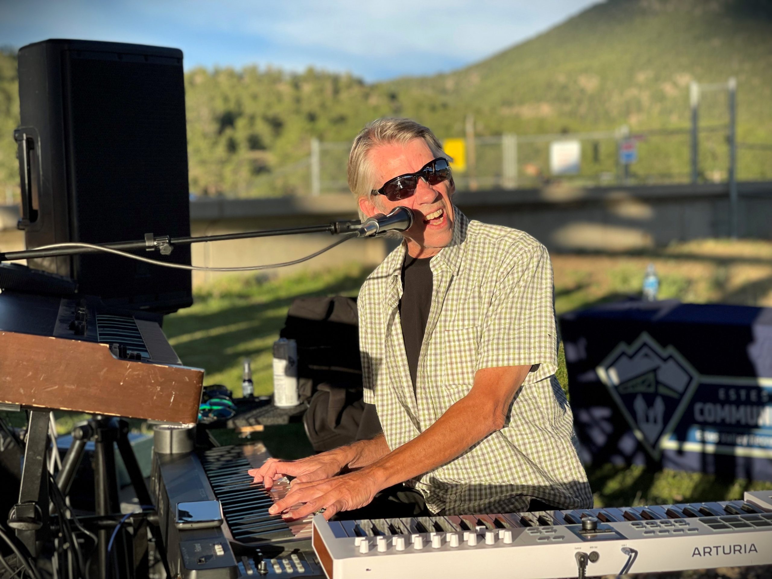 An older adult is playing keyboard outdoors, smiling and singing into a microphone. Trees and a mountain are in the background.