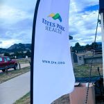 A vertical flag with "Estes Park Health" and a logo printed on it, standing outdoors next to a parking area with a red truck and mountains in the background. The flag also displays the URL "www.eph.org".