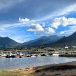 A marina with several boats docked on a calm lake, with mountain ranges and a partly cloudy sky in the background.