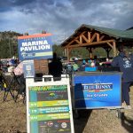 Outdoor entrance area of Lake Estes Marina Pavilion, featuring event signs, a Blue Bunny ice cream sign, and people gathered under a wooden pavilion. The sky is partly cloudy.