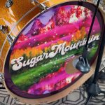 Close-up of a bass drum with a "Sugar Mountain" logo and scenic imagery printed on the drumhead. The drum is set up on a patterned rug with a microphone positioned nearby.