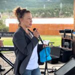 A woman holding a microphone speaks outdoors while another woman stands nearby. Musical equipment, including a keyboard and speakers, are set up behind them.