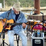 An older man wearing a cap and denim clothes plays an acoustic guitar on stage. There is a drum set and various music equipment in the background.